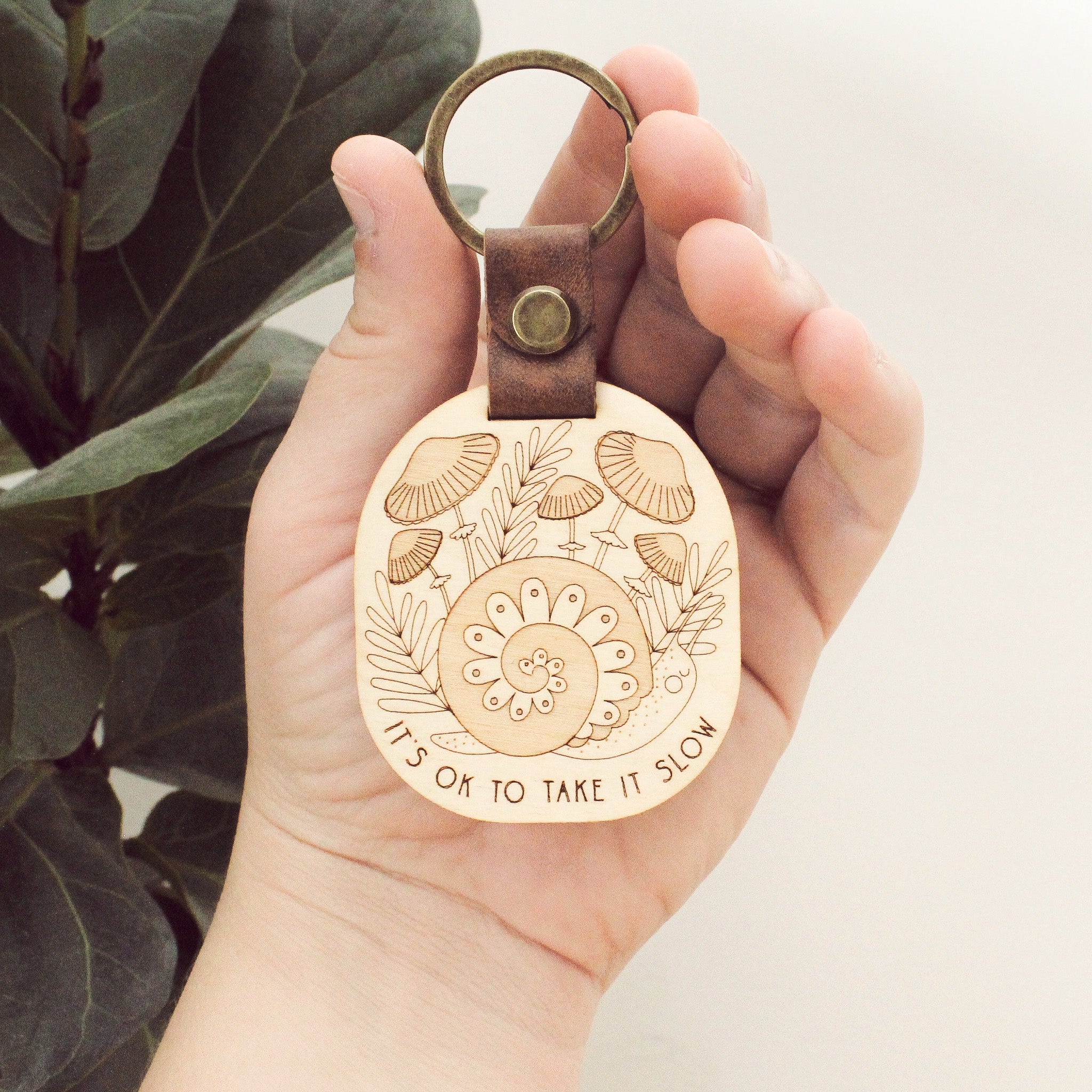wooden keychain with snail design surrounded by mushrooms and the text 'it's ok to take it slow.' with leather strap and bronze colored details.