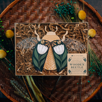 load image into gallery viewer, decorative wooden beetle, laser cut and layered with etched base, clear acrylic wings with a leaf design, and floral inlaid shell. wings and shell are movable as they&#39;re connected with a brass fastener. shell piece is black with a white flower with green leaves. shown packaged in a box with kraft paper strips.
