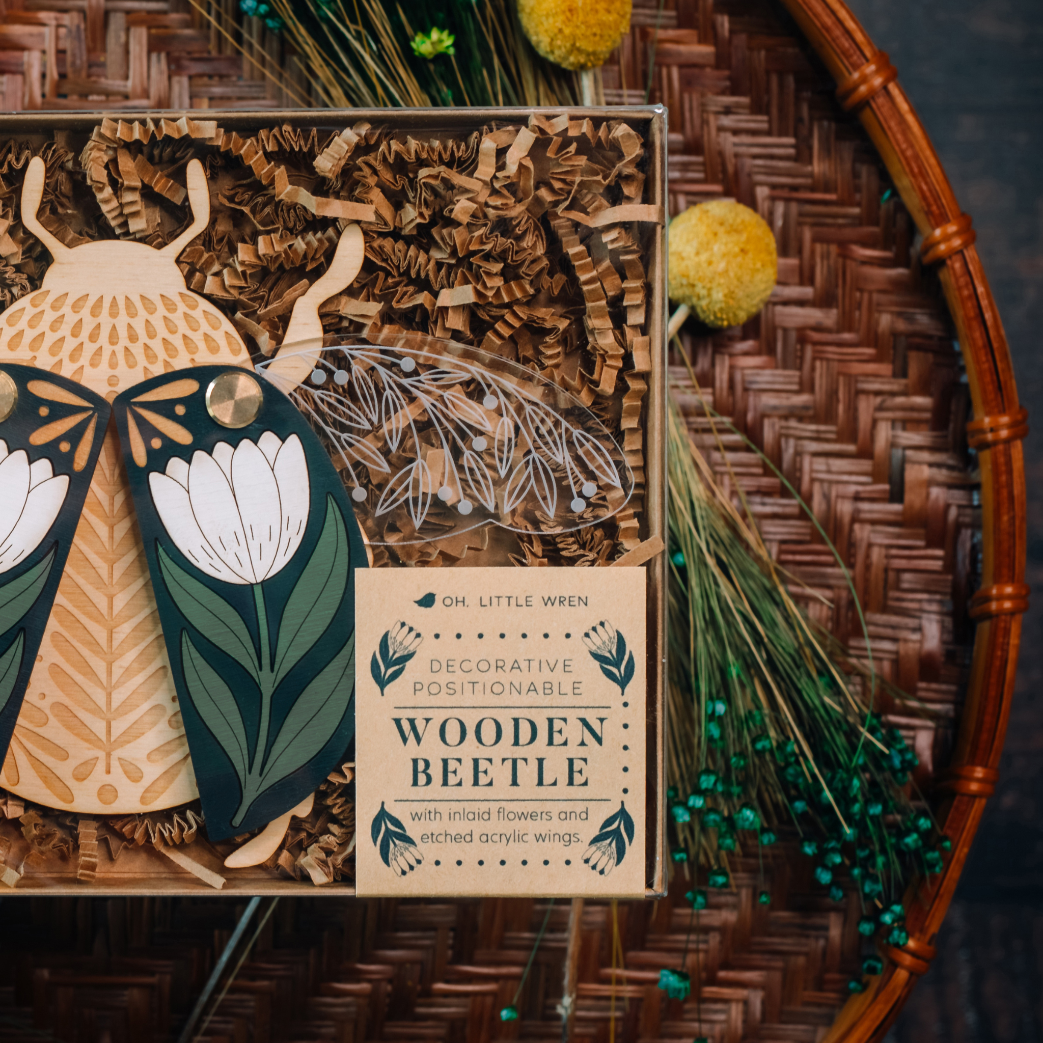 decorative wooden beetle, laser cut and layered with etched base, clear acrylic wings with a leaf design, and floral inlaid shell. wings and shell are movable as they're connected with a brass fastener. shell piece is black with a white flower with green leaves. shown closeup, packaged in a box with kraft paper strips.