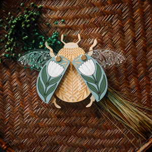 decorative wooden beetle, laser cut and layered with etched base, clear acrylic wings with a leaf design, and floral inlaid shell. wings and shell are movable as they're connected with a brass fastener. shell piece is dusty blue with a white flower with green leaves.