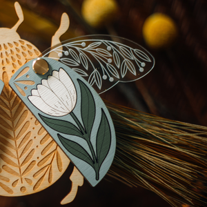 decorative wooden beetle, laser cut and layered with etched base, clear acrylic wings with a leaf design, and floral inlaid shell. wings and shell are movable as they're connected with a brass fastener. shell piece is dusty blue with a white flower with green leaves. shown close up of right half.