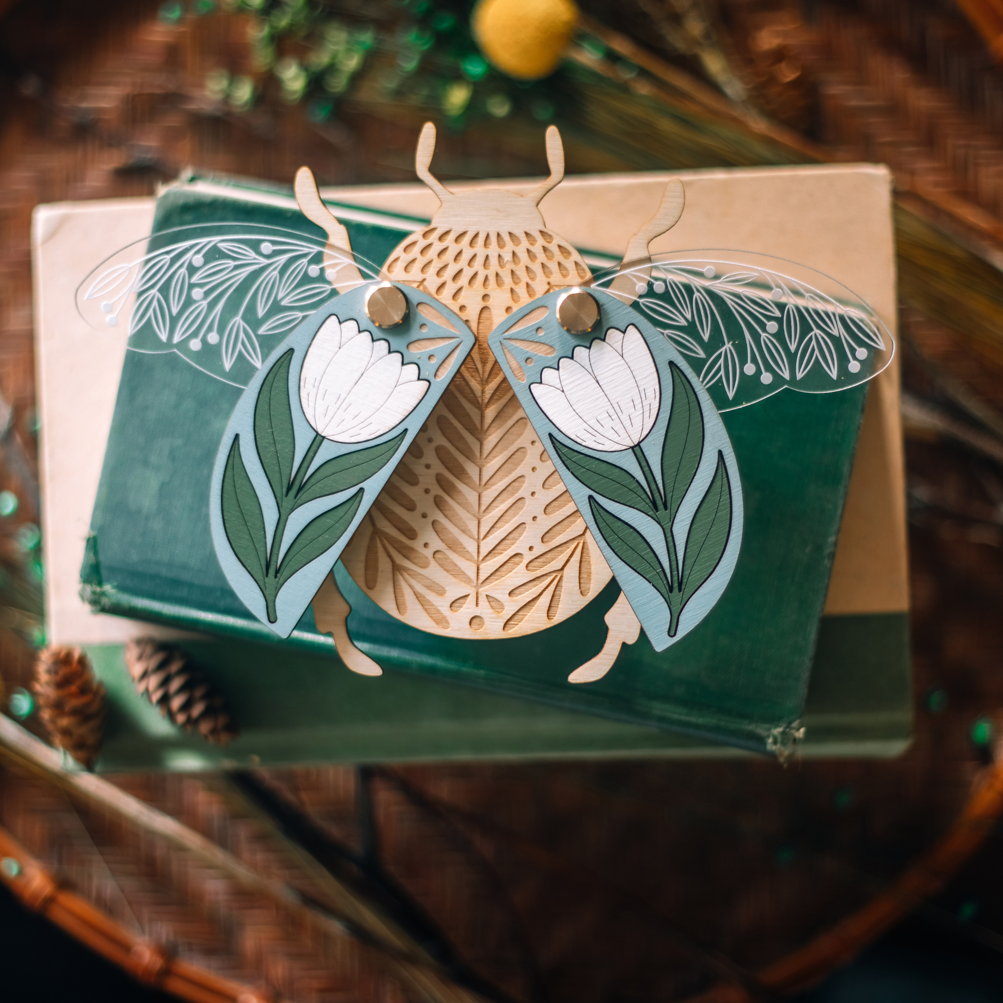 decorative wooden beetle, laser cut and layered with etched base, clear acrylic wings with a leaf design, and floral inlaid shell. wings and shell are movable as they're connected with a brass fastener. shell piece is dusty blue with a white flower with green leaves. shown staged on a stack of books.