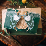 load image into gallery viewer, decorative wooden beetle, laser cut and layered with etched base, clear acrylic wings with a leaf design, and floral inlaid shell. wings and shell are movable as they&#39;re connected with a brass fastener. shell piece is dusty blue with a white flower with green leaves. shown staged on a stack of books.
