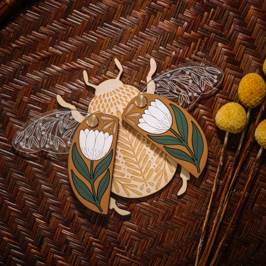 decorative wooden beetle, laser cut and layered with etched base, clear acrylic wings with a leaf design, and floral inlaid shell. wings and shell are movable as they're connected with a brass fastener. shell piece is mustard yellow with a white flower with green leaves.