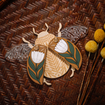 load image into gallery viewer, decorative wooden beetle, laser cut and layered with etched base, clear acrylic wings with a leaf design, and floral inlaid shell. wings and shell are movable as they&#39;re connected with a brass fastener. shell piece is mustard yellow with a white flower with green leaves.
