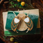load image into gallery viewer, decorative wooden beetle, laser cut and layered with etched base, clear acrylic wings with a leaf design, and floral inlaid shell. wings and shell are movable as they&#39;re connected with a brass fastener. shell piece is mustard yellow with a white flower with green leaves. shown staged on a stack of books.
