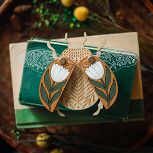decorative wooden beetle, laser cut and layered with etched base, clear acrylic wings with a leaf design, and floral inlaid shell. wings and shell are movable as they're connected with a brass fastener. shell piece is mustard yellow with a white flower with green leaves. shown staged on a stack of books.