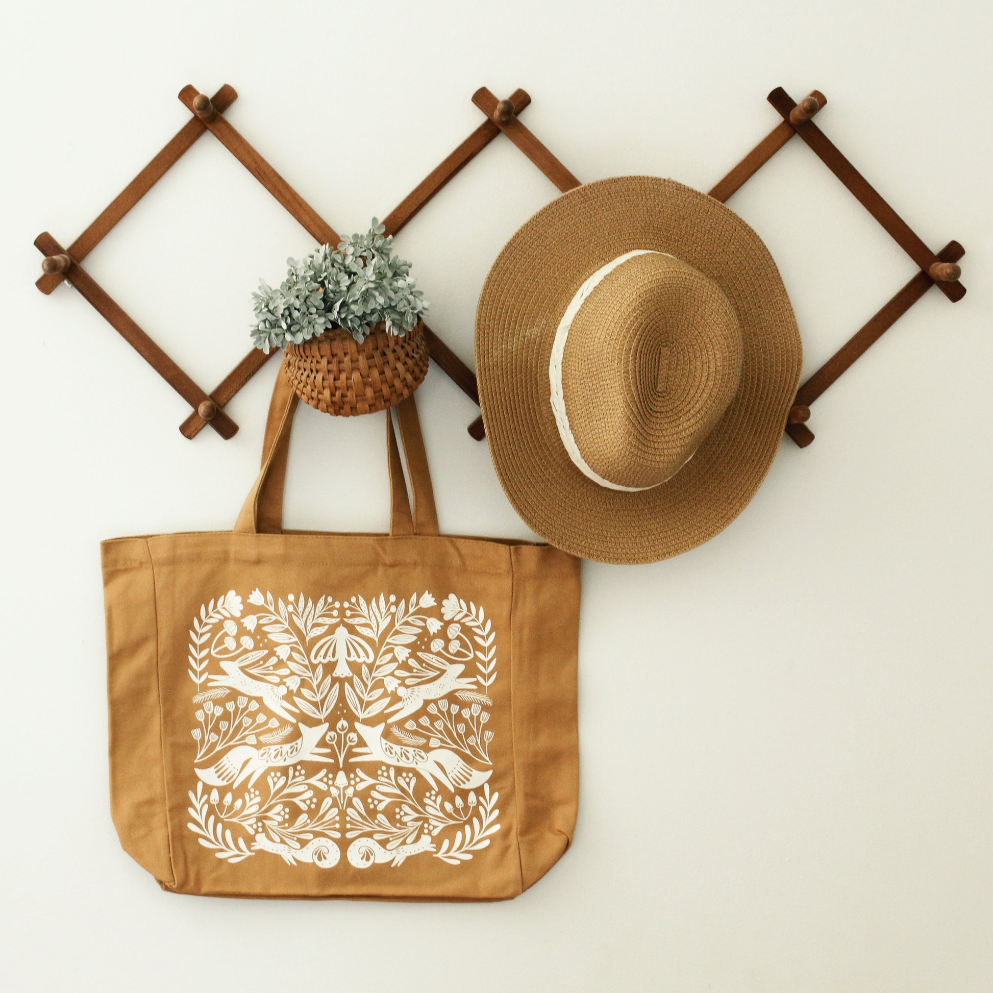gusseted tote bag in burnt orange with a detailed silk screen design in warm white with florals, mushrooms, foxes, squirrels, rabbits and a bird. shown staged hanging on a wood accordion rack with a straw hat and greenery.