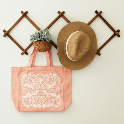 gusseted tote bag in a coral pink with a detailed silk screen design in warm white with florals, mushrooms, foxes, squirrels, rabbits and a bird. shown staged hanging on a wood accordion rack with a straw hat and greenery.