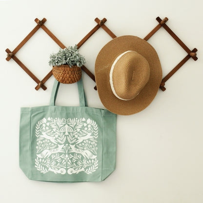 gusseted tote bag in ocean blue with a detailed silk screen design in warm white with florals, mushrooms, foxes, squirrels, rabbits and a bird. shown staged hanging on a wood accordion rack with a straw hat and greenery.