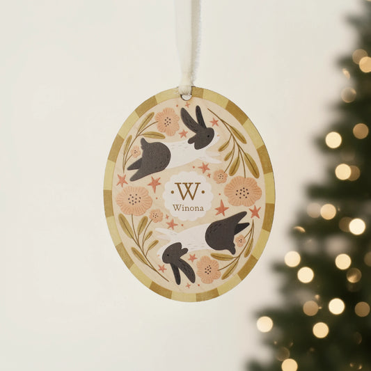 Illustrated wooden ornament with rabbits and flowers and the center monogram 'W' with 'Winona' text below, hanging against a blurred Christmas tree background. Ornament colors are yellows, pinks, black, and white. 