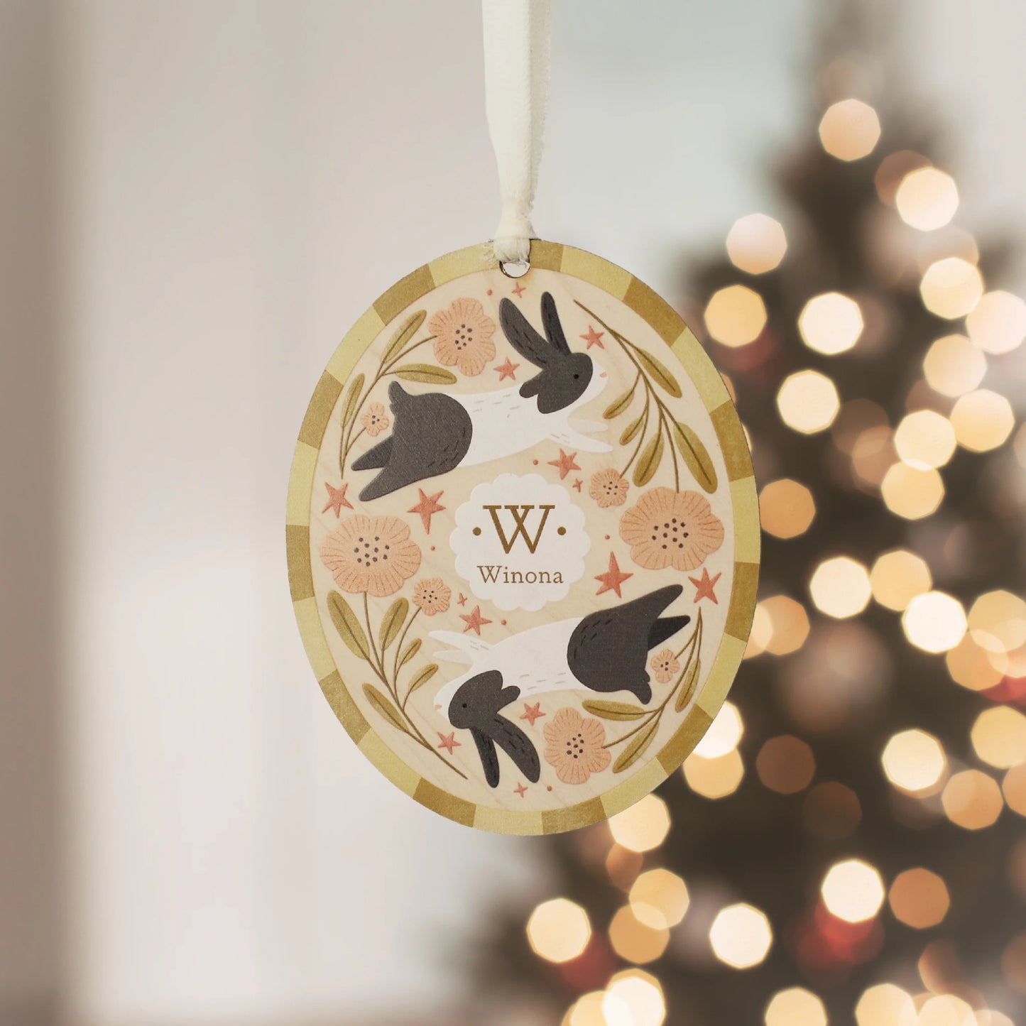Illustrated wooden ornament with rabbits and flowers and the center monogram 'W' with 'Winona' text below, hanging against a blurred Christmas tree background. Ornament colors are yellows, pinks, black, and white. 