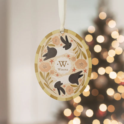Illustrated wooden ornament with rabbits and flowers and the center monogram 'W' with 'Winona' text below, hanging against a blurred Christmas tree background. Ornament colors are yellows, pinks, black, and white. 