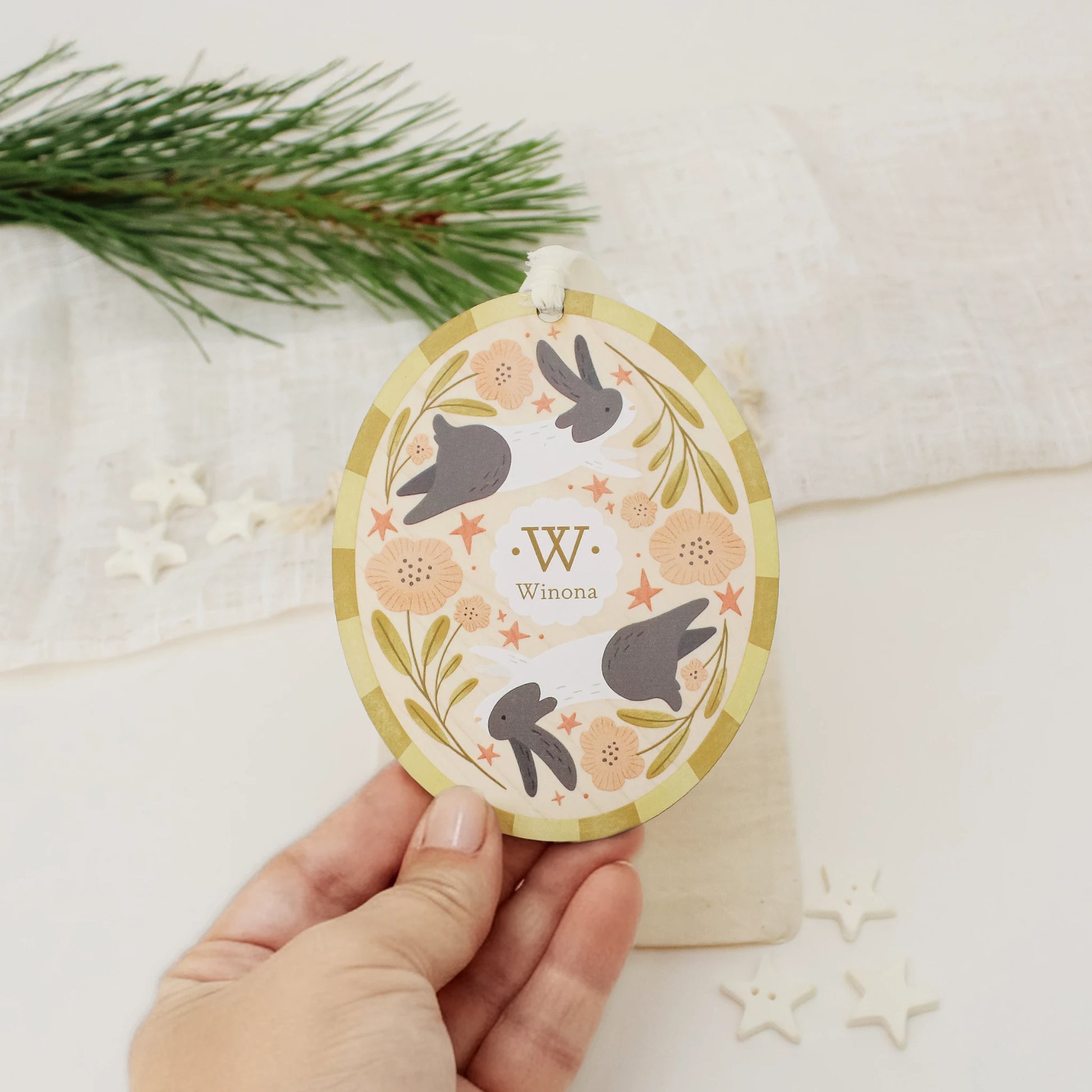 Illustrated wooden ornament with rabbits and flowers and the center monogram 'W' with 'Winona' text below, held by a hand against a light background. Ornament colors are yellows, pinks, black, and white. 