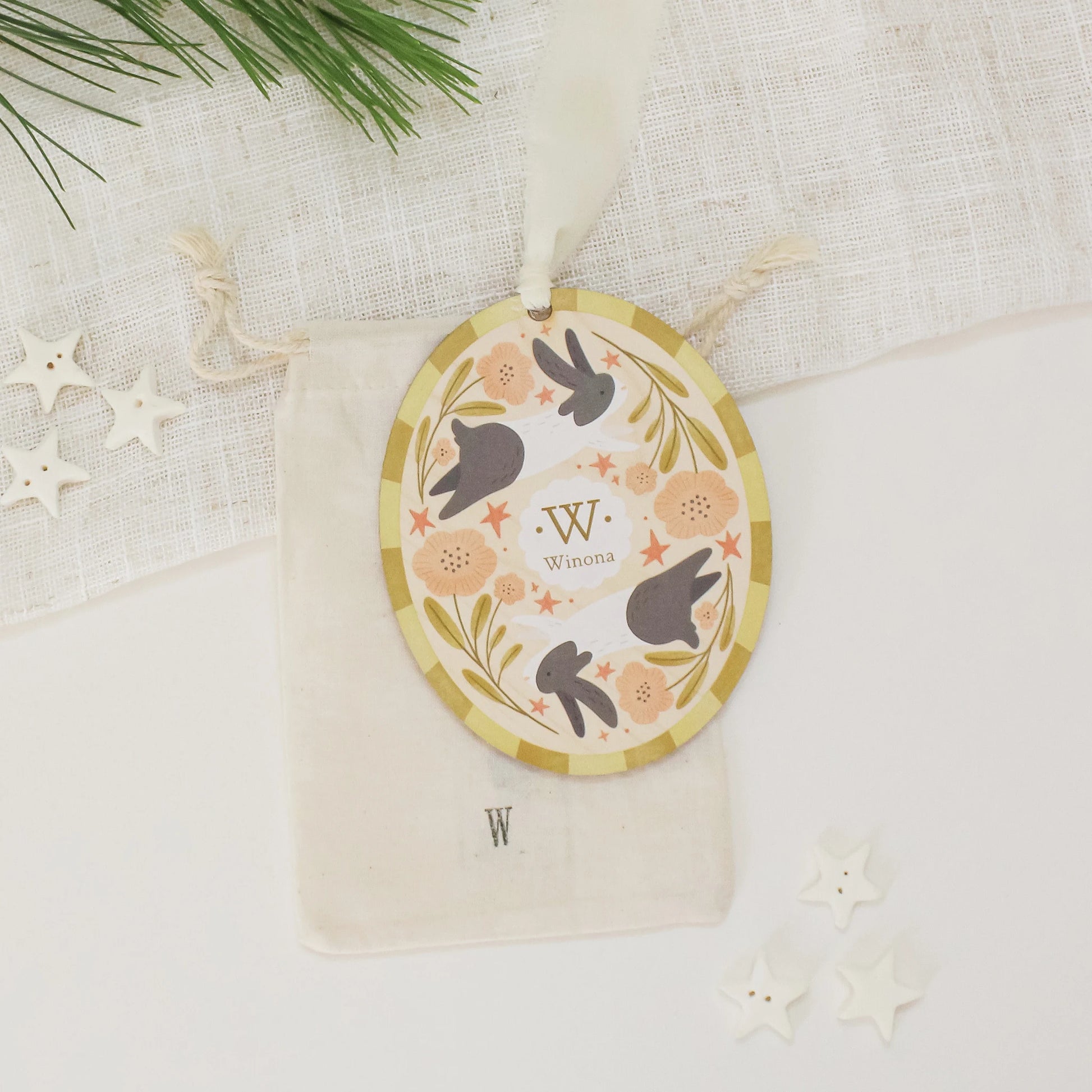 Illustrated wooden ornament with rabbits and flowers and the center monogram 'W' with 'Winona' text below, lying on a cotton bag stamped with the letter 'W' against a light background. Ornament colors are yellows, pinks, black, and white. 