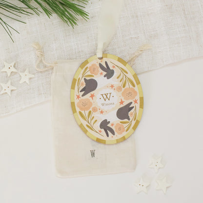 Illustrated wooden ornament with rabbits and flowers and the center monogram 'W' with 'Winona' text below, lying on a cotton bag stamped with the letter 'W' against a light background. Ornament colors are yellows, pinks, black, and white. 