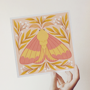art print showing a rosy maple moth in bright pinks and yellows, surrounded by yellow and white whimsical florals. shown being held in a hand.