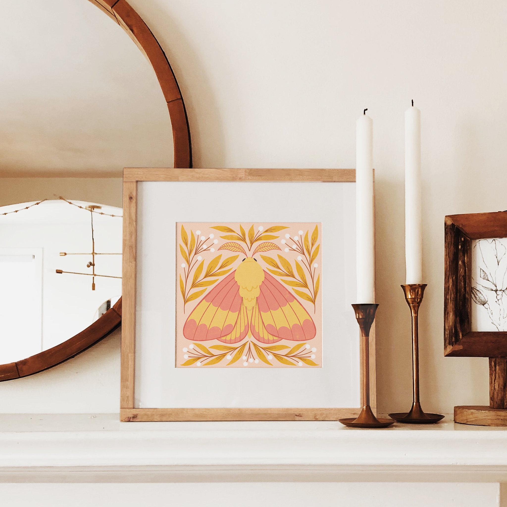 art print showing a rosy maple moth in bright pinks and yellows, surrounded by yellow and white whimsical florals. shown displayed in a wood frame on a mantle.