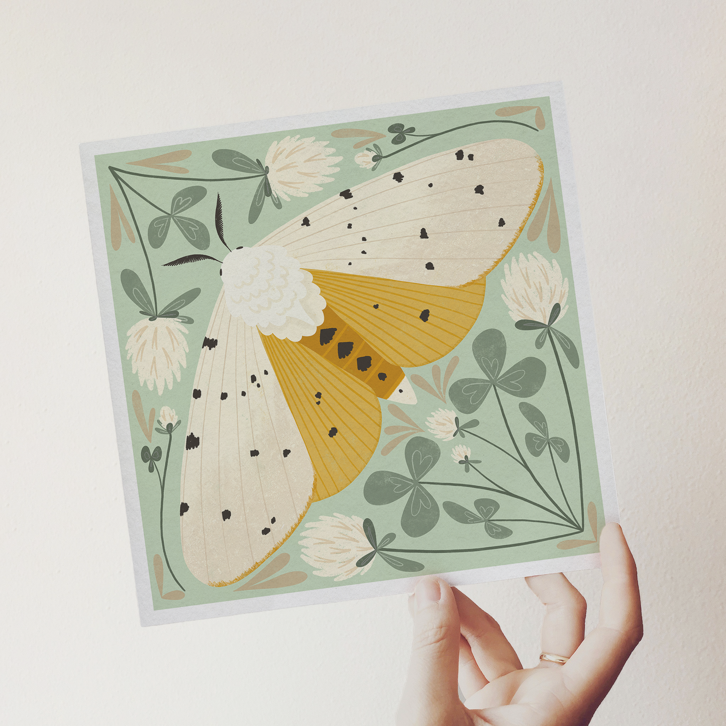 art print showing a salt marsh moth in black, white and mustard yellow, surrounded by whimsical mint green clovers and white clover flowers. shown being held in a hand.