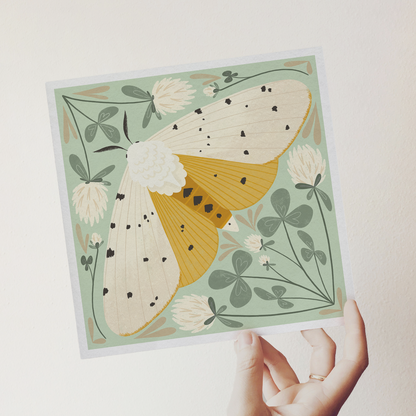 art print showing a salt marsh moth in black, white and mustard yellow, surrounded by whimsical mint green clovers and white clover flowers. shown being held in a hand.