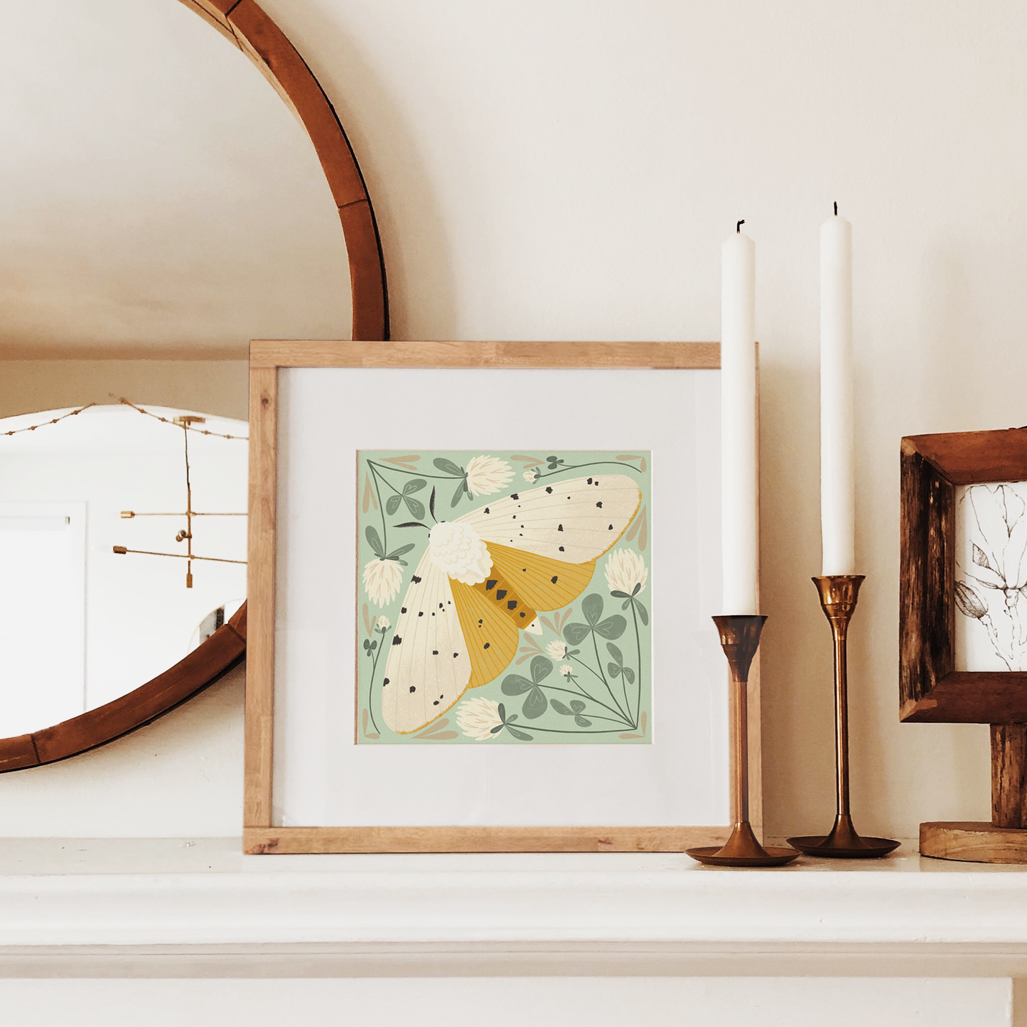 art print showing a salt marsh moth in black, white and mustard yellow, surrounded by whimsical mint green clovers and white clover flowers. shown displayed in a wood frame, resting on a mantle.
