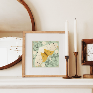 art print showing a salt marsh moth in black, white and mustard yellow, surrounded by whimsical mint green clovers and white clover flowers. shown displayed in a wood frame, resting on a mantle.