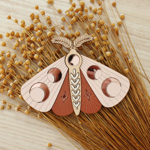 wooden moth with moon phase design. painted in blush pinks and terracotta reds with wood tones and mirrored rose gold moon phase inlays.