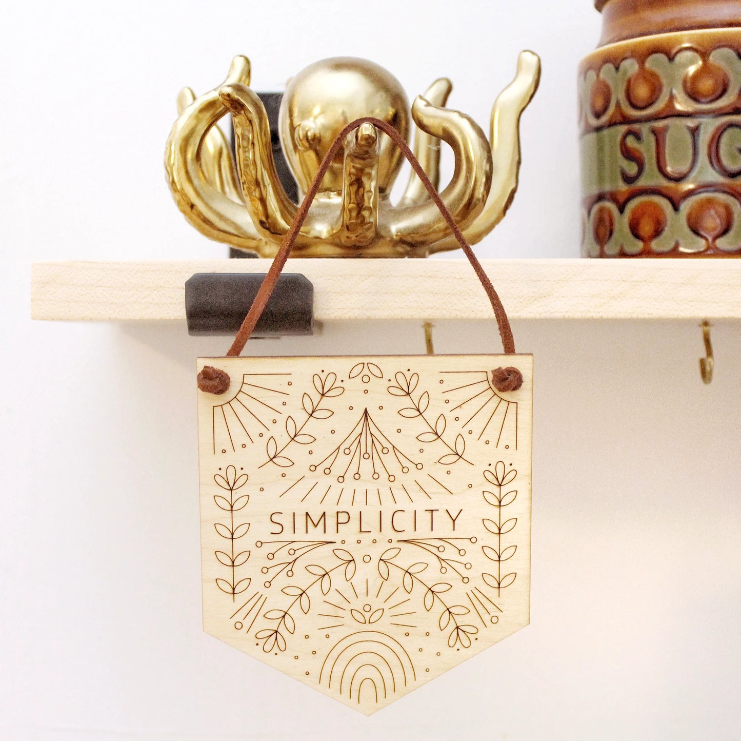 word of the year mini banner hanging from a shelf the word SIMPLICITY surrounded by intricately etched designs of flowers, rainbows and other motifs.