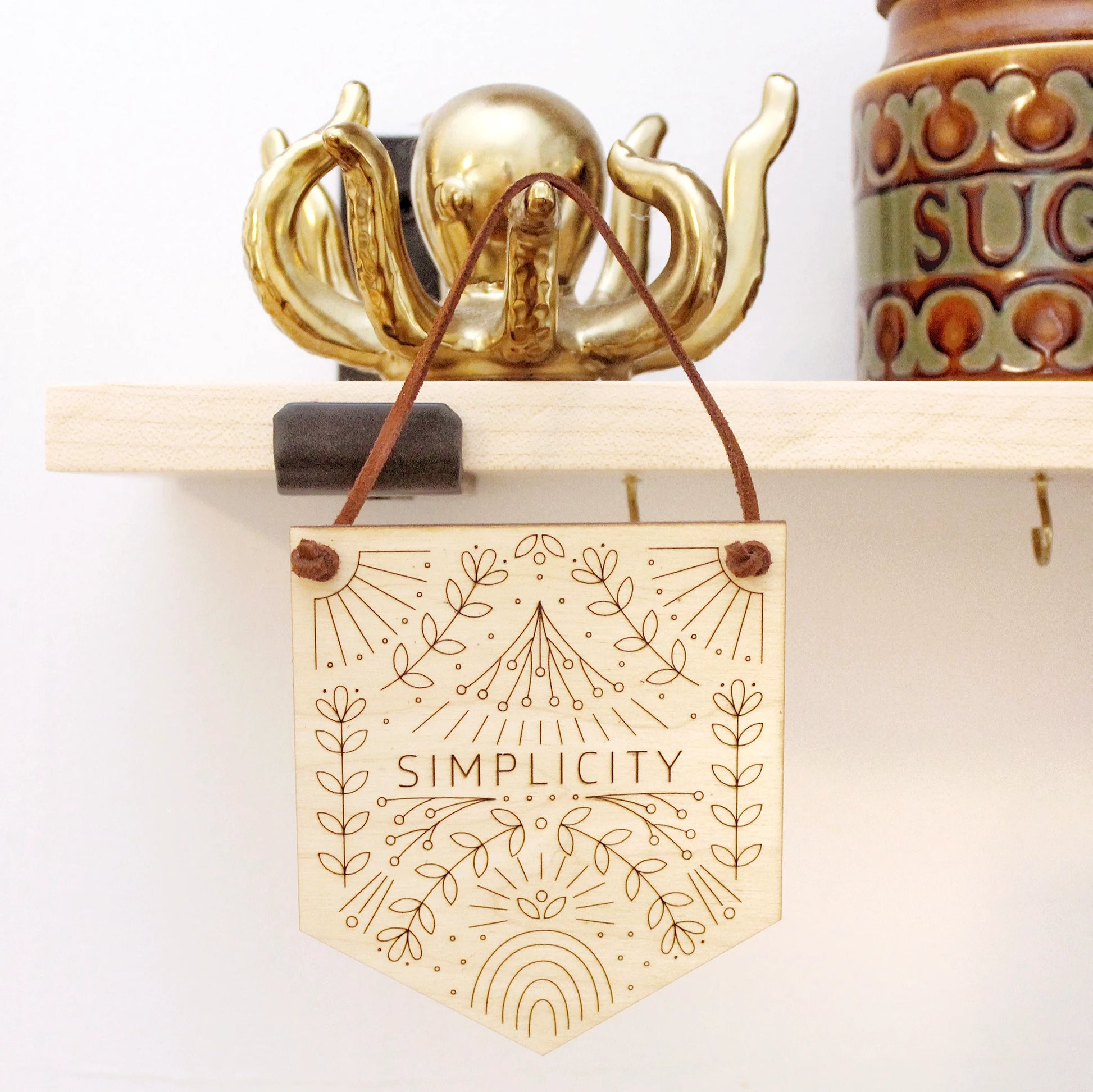 word of the year mini banner hanging from a shelf the word SIMPLICITY surrounded by intricately etched designs of flowers, rainbows and other motifs.