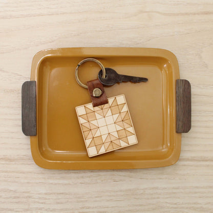 wooden keychain with quilt block pattern in two wood tones, with leather strap and bronze colored details.