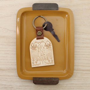 wooden keychain for the adventure lover. design has mountains, trees, fields and streams and the words 'adventure awaits'.  wooden keychain with leather strap and bronze colored hardware.
