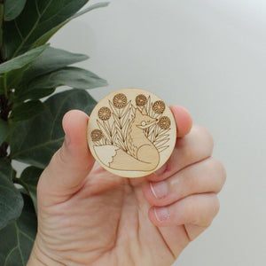pair of circular wooden magnets, one with a fox surrounded by flowers and the other with a snail and mushrooms.